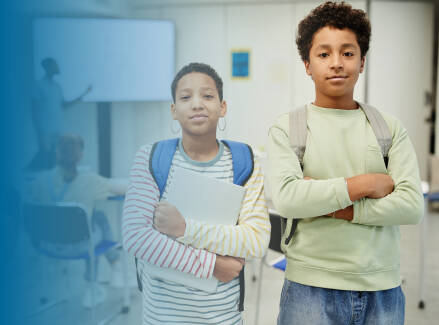 Two students in a classroom wearing backpacks and holding notebooks.